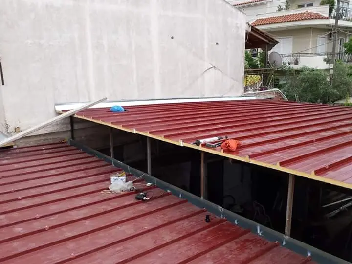 Μερικώς κατασκευασμένη στέγη με κόκκινες λαμαρίνες και πανελ πολυουρεθάνησ. Διάφορα εργαλεία και υλικά είναι διάσπαρτα στην κορυφή, όπως ένα μπλε καπάκι και ένα πορτοκαλί γάντι. Το φόντο δείχνει έναν λευκό τοίχο και λίγο πράσινο.