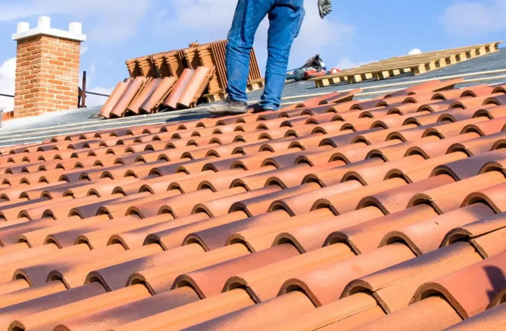 Ένα άτομο που φορά μπλε τζιν εργάζεται σε μια κόκκινη κεραμοσκεπή, που περιβάλλεται από πήλινα πλακάκια και διάσπαρτα γύρω τους πάνελ πολυουρεθάνης. Μια καμινάδα υπάρχει καθώς και ο καταγάλανος ουρανός.