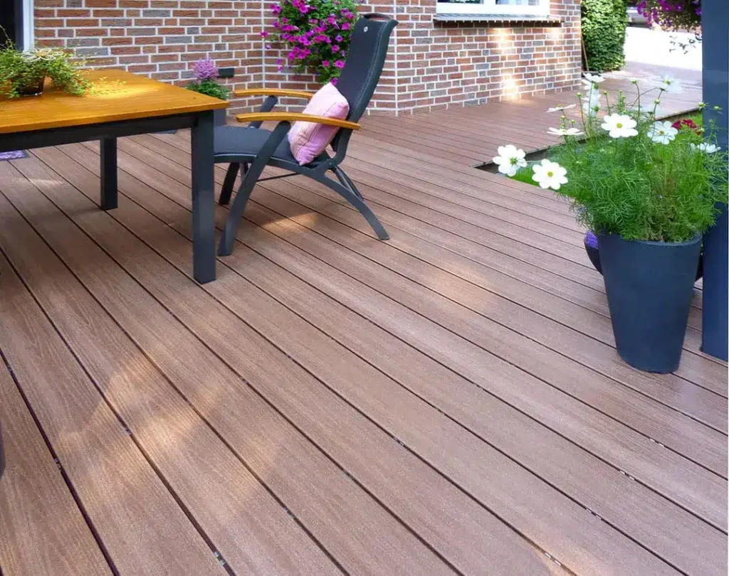 ντεκ βεράντας με όψη ξύλου που είναι κατασκευασμένο από συνθετικό wpc υλικό με τραπέζι και καρέκλα. Το τραπέζι έχει φυτό σε γλάστρα και η καρέκλα έχει ροζ μαξιλάρι. Γλάστρες με λευκά και ροζ άνθη βρίσκονται σε κοντινή απόσταση. Το φόντο διαθέτει τοίχο από κόκκινο τούβλο και περισσότερα φυτά.