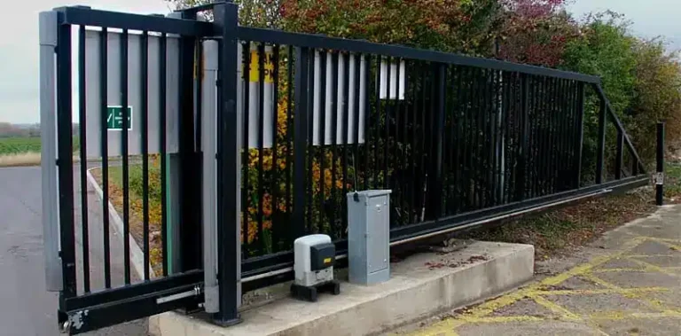 Μια μαύρη μεταλλική συρόμενη πύλη με κάθετες ράβδους απλώνεται σε όλο το δρόμο, ενισχυμένη από έναν αυτοματοποιημένο μηχανισμό συρόμενης πόρτας στη βάση της. Είναι όμορφα πλαισιωμένο από γύρω πράσινο και οδικές σημάνσεις.